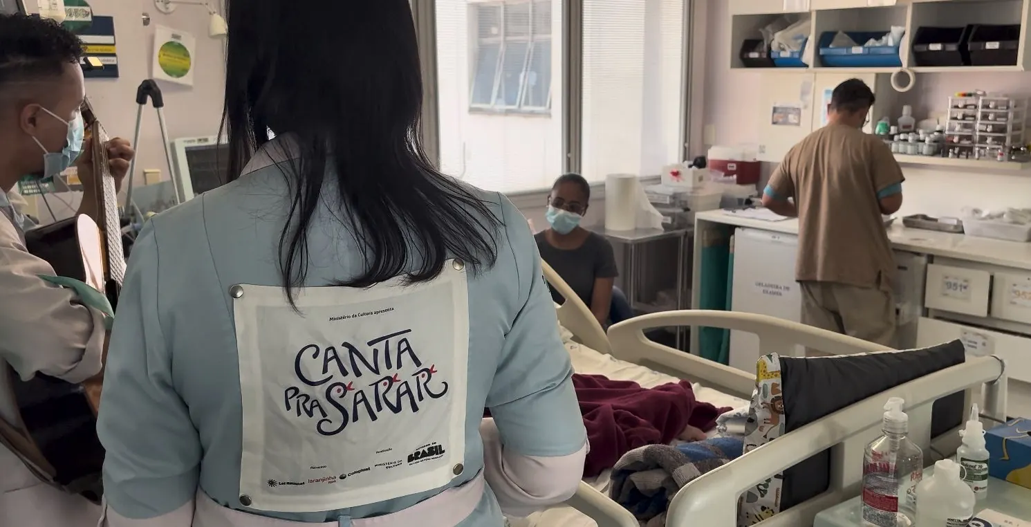 Artista de costas, com o jaleco do Canta Pra Sarar, diante de uma acompanhante no quarto