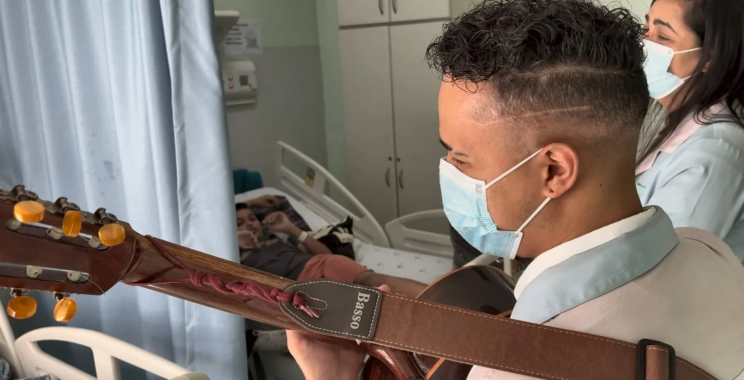 Artista toca violão para um paciente deitado, num quarto do Hospital São Paulo
