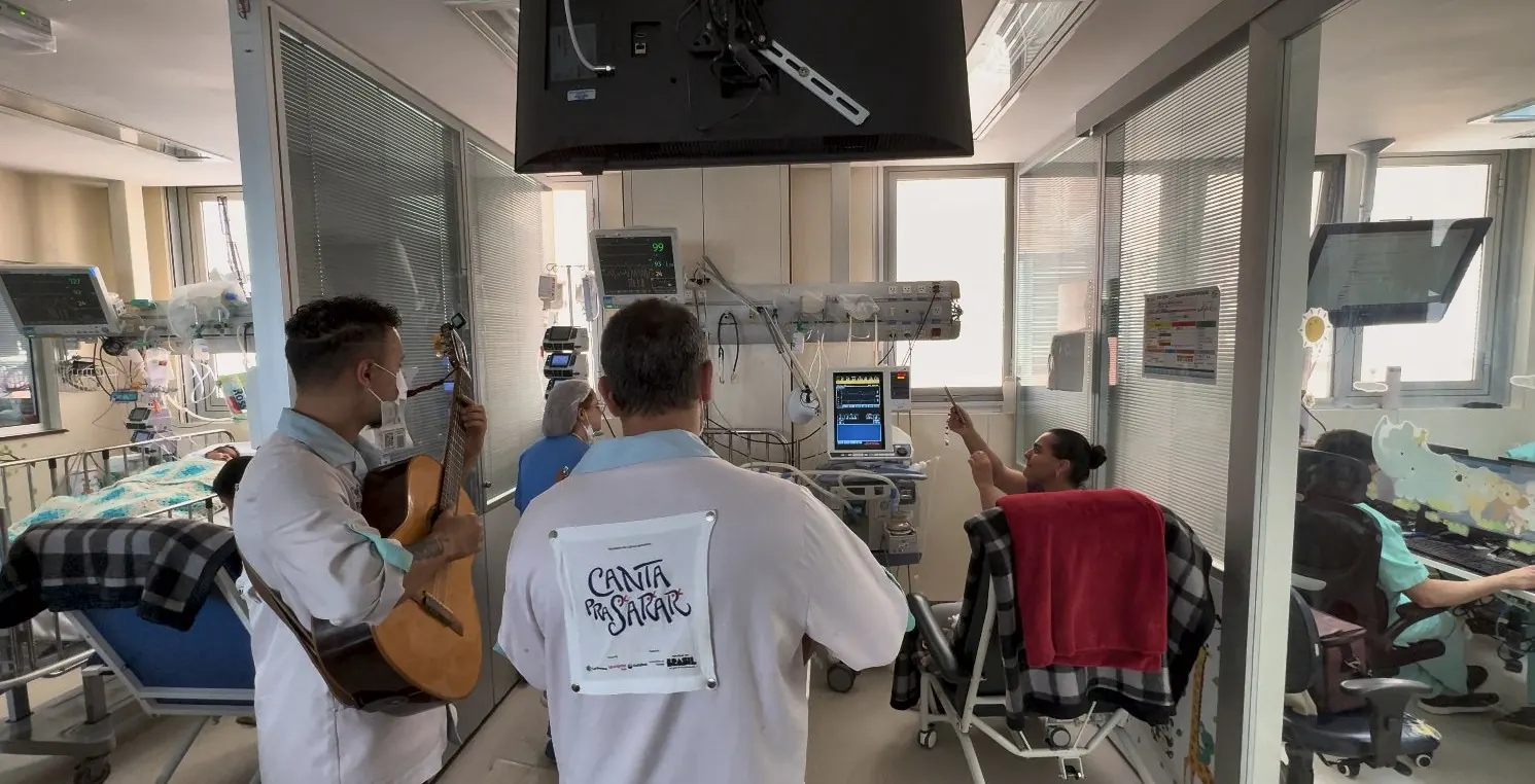 Artistas tocam para uma paciente sentada na poltrona, ao lado do leito