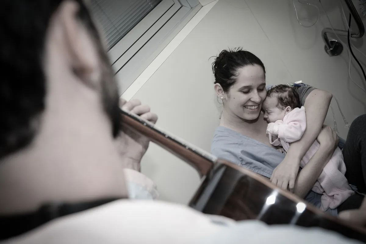 Artista do Saracura toca violão para uma mãe que segura o bebê no colo, num quarto de hospital